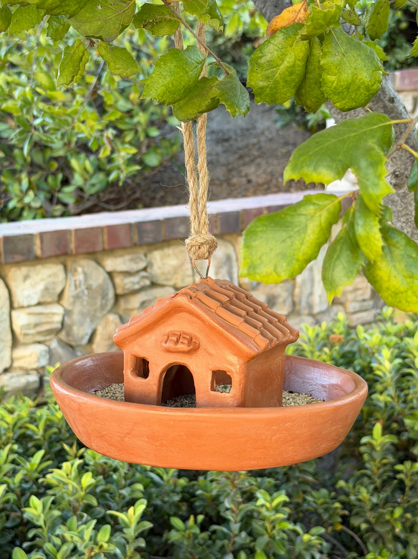 A terracotta clay bird feeder shaped like a small house, suspended by a rope from a tree branch over a garden hedge.